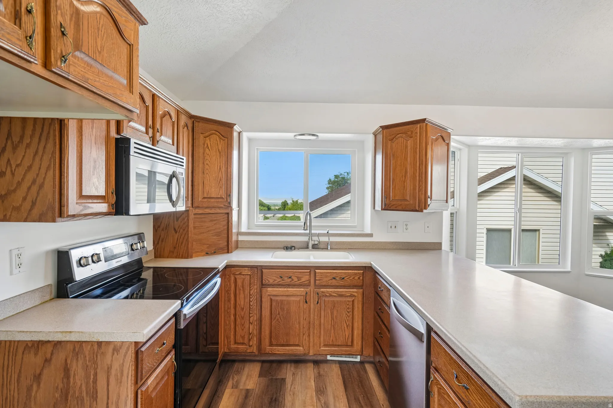 Kitchen featuring wood cabinetry, light countertops, wood-finish flooring, stainless steel appliances, and a double basin sink beneath a window