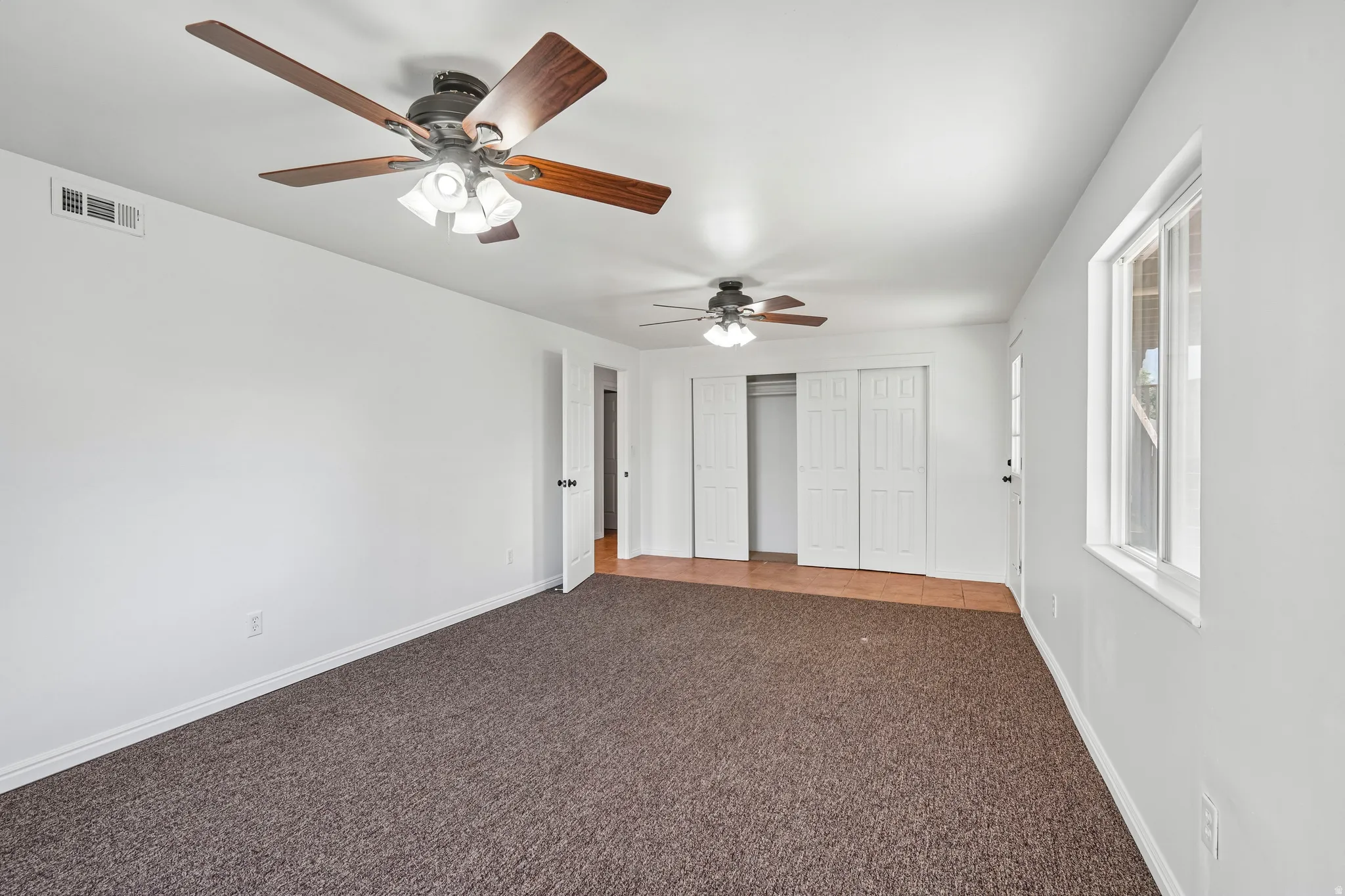 Spacious room featuring brown carpeting, a large window, a ceiling fan with integrated lighting, and white bi-fold closet doors