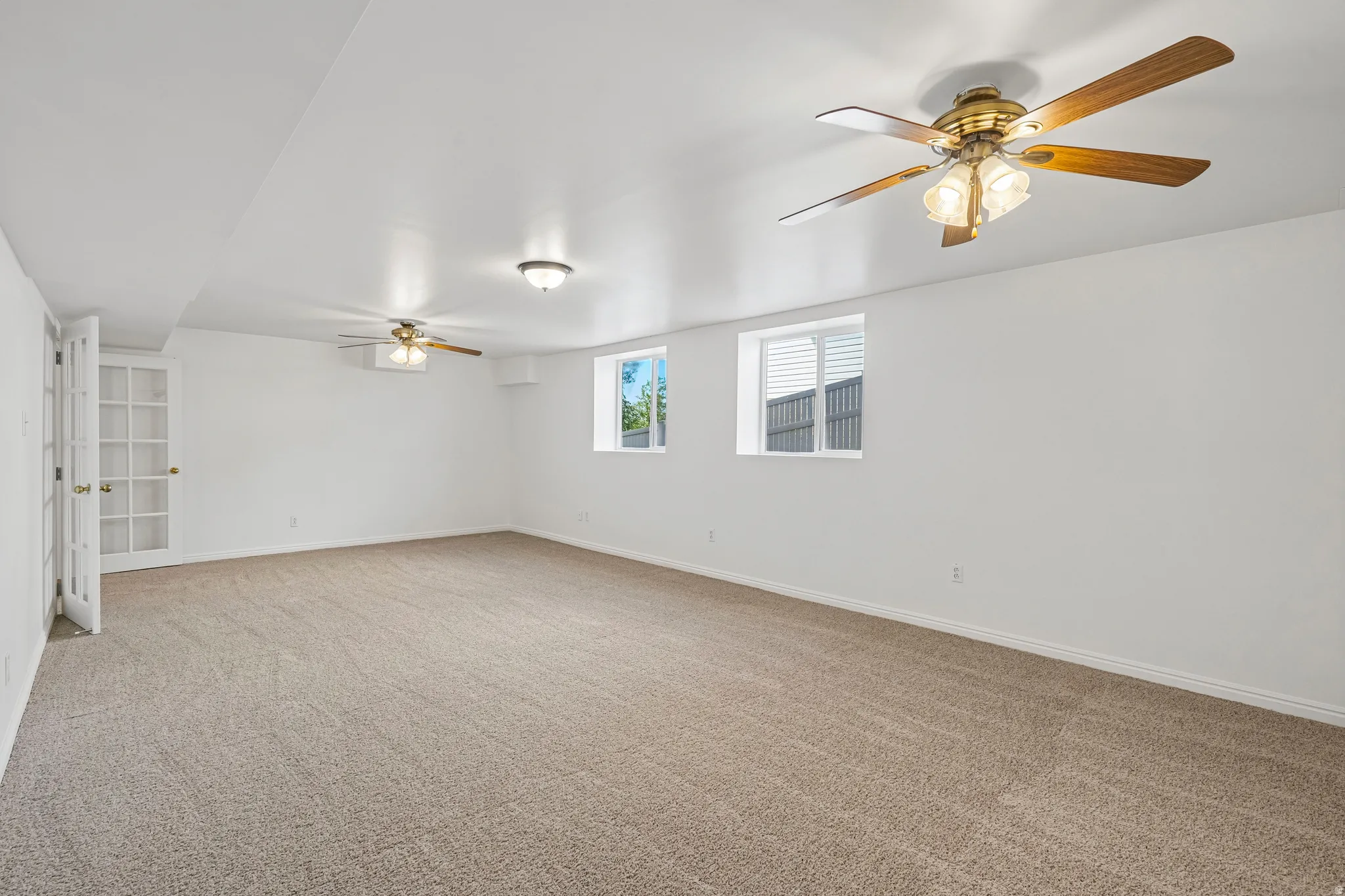 Expansive carpeted room featuring two brass-finish ceiling fans with wood-finish blades, two recessed windows with blinds, and double French doors