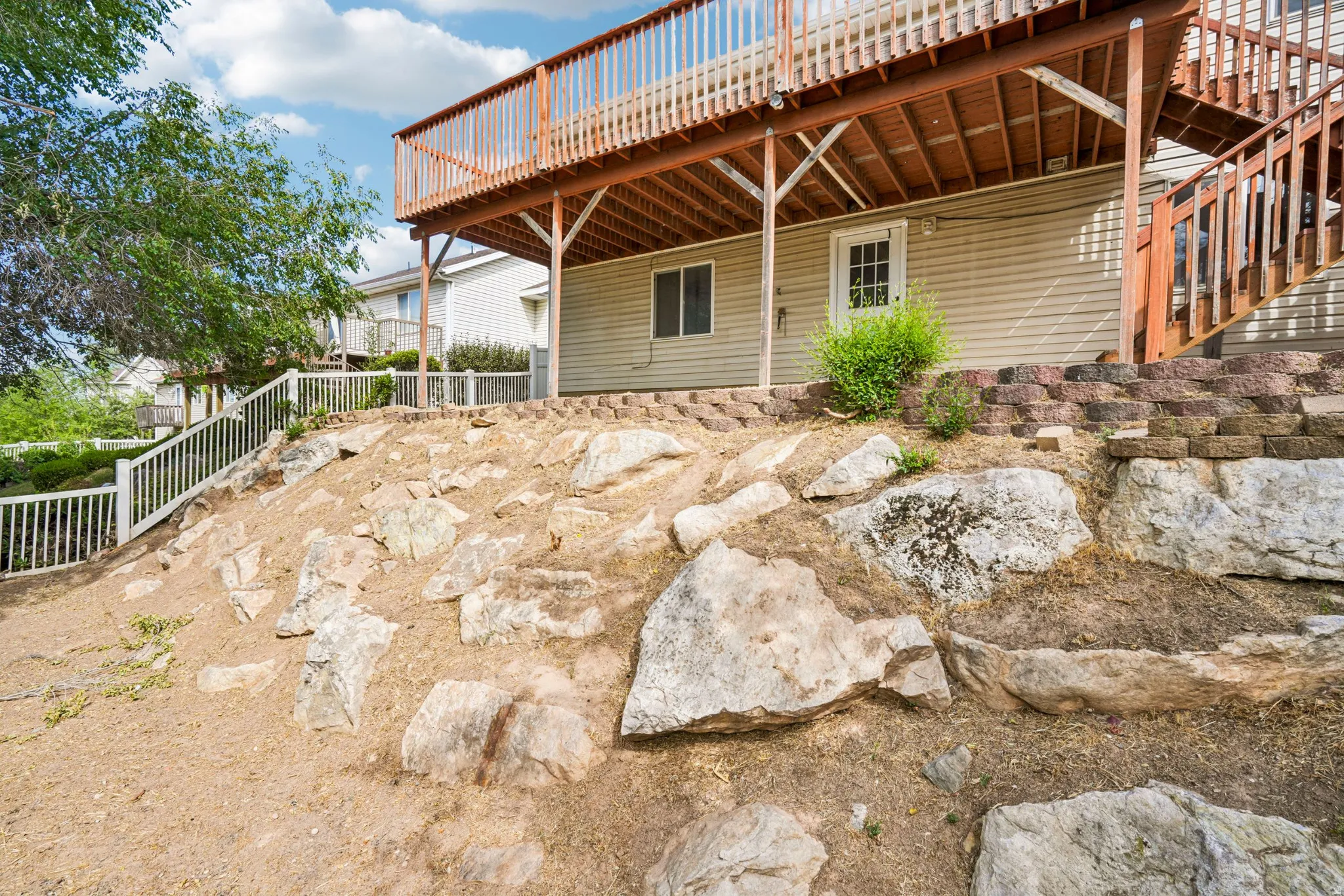 Multi-level backyard featuring a natural rock landscaping on a sloped grade, a retaining wall constructed from concrete blocks, a walk-out basement entry, and an upper-level wood deck with matching wood stairs and railing