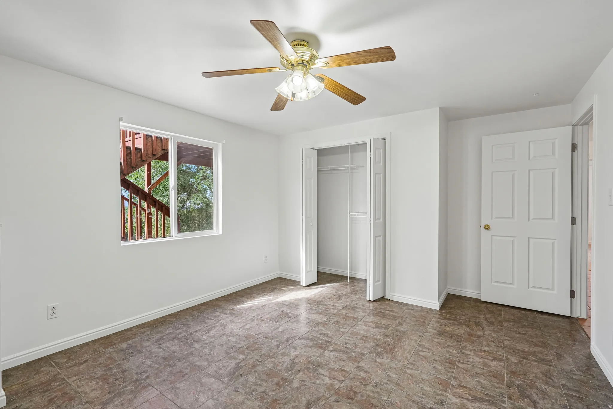Bright room featuring neutral-toned tile flooring, a window with a treed outdoor outlook, a ceiling fan with integrated lighting, a bi-fold closet, and a paneled entry door