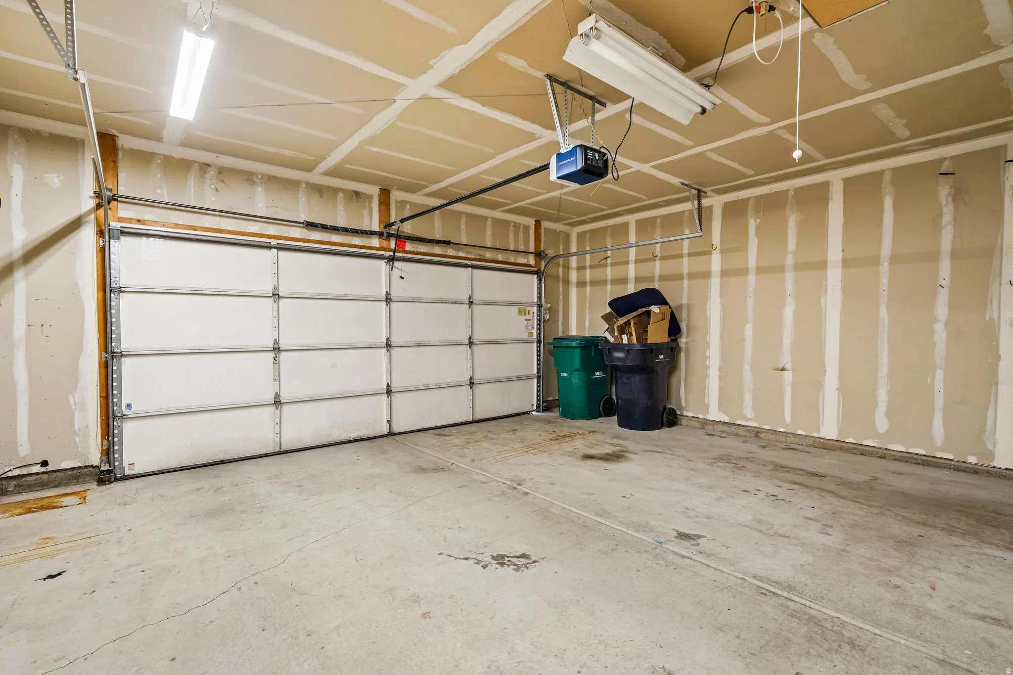 Spacious garage featuring a concrete floor, exposed stud walls, overhead fluorescent lighting, and a white panel garage door with an automatic opener