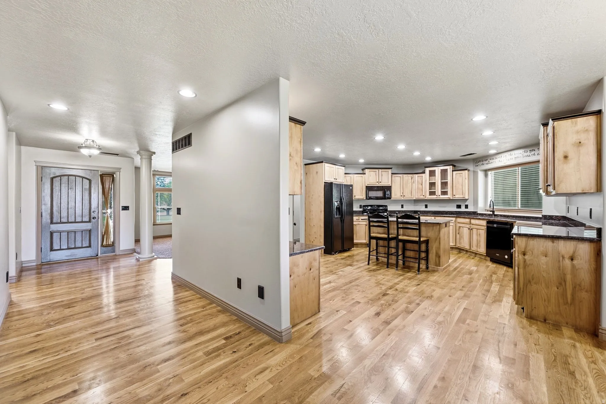 Open-concept kitchen featuring light wood cabinetry, dark countertops, black appliances, and a central island with seating