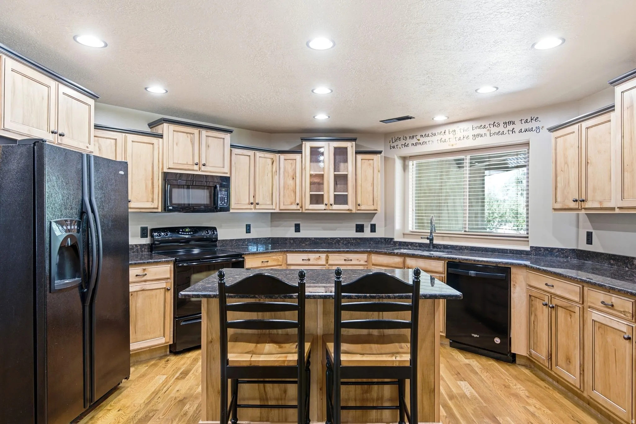 Spacious kitchen featuring light wood cabinetry with dark trim, dark granite-style countertops, wood-finish flooring, and a central island with seating