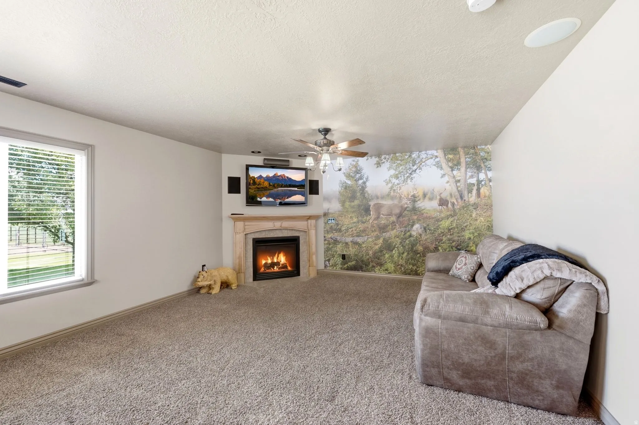 Living area featuring a corner fireplace with a stone-finish mantel, wall-mounted television, ceiling fan with integrated lighting, and light-toned carpeting