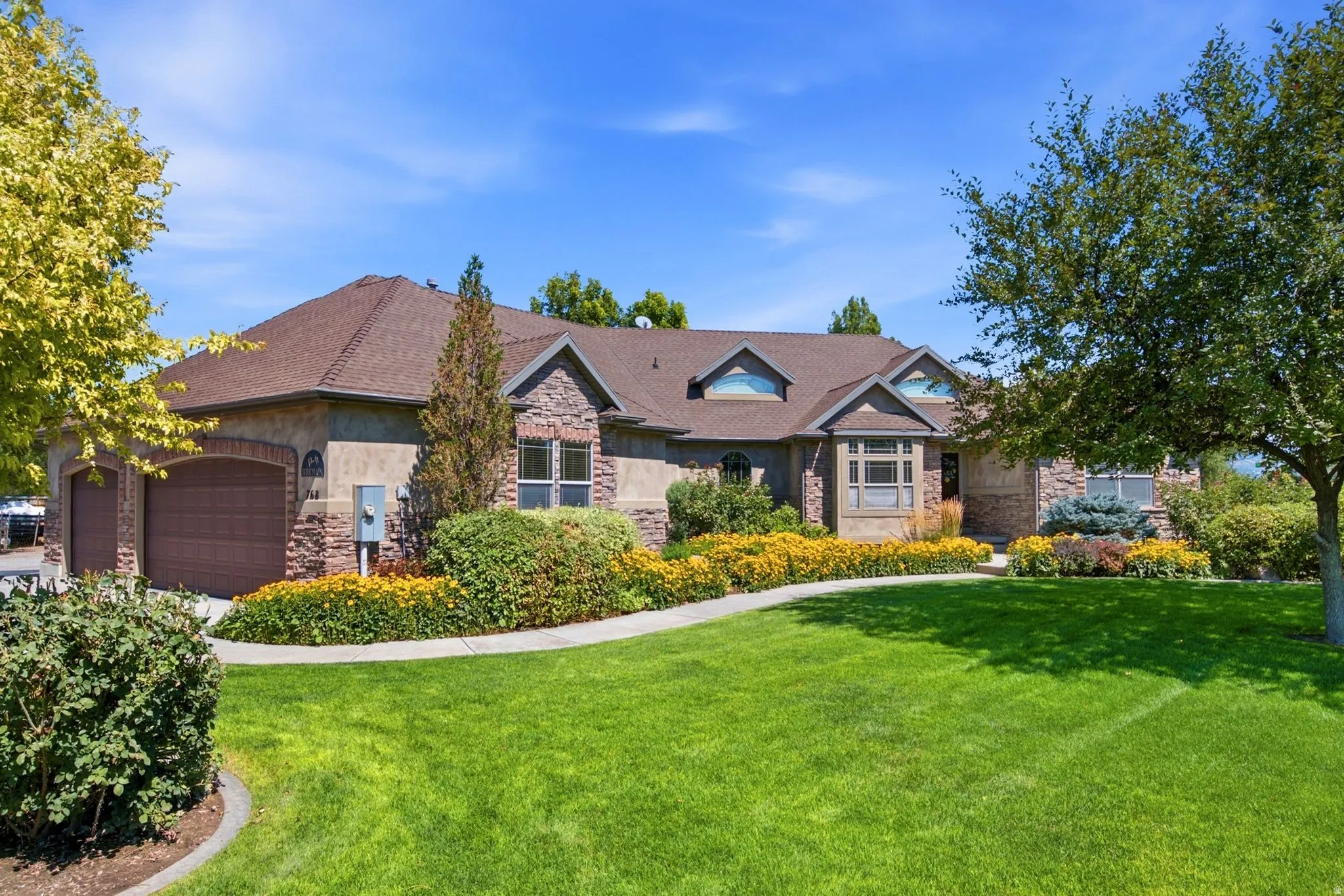 Craftsman-style home featuring a stone and stucco exterior, arched double garage doors, a bay window, a gabled roofline, and established landscaping with mature trees and flower beds