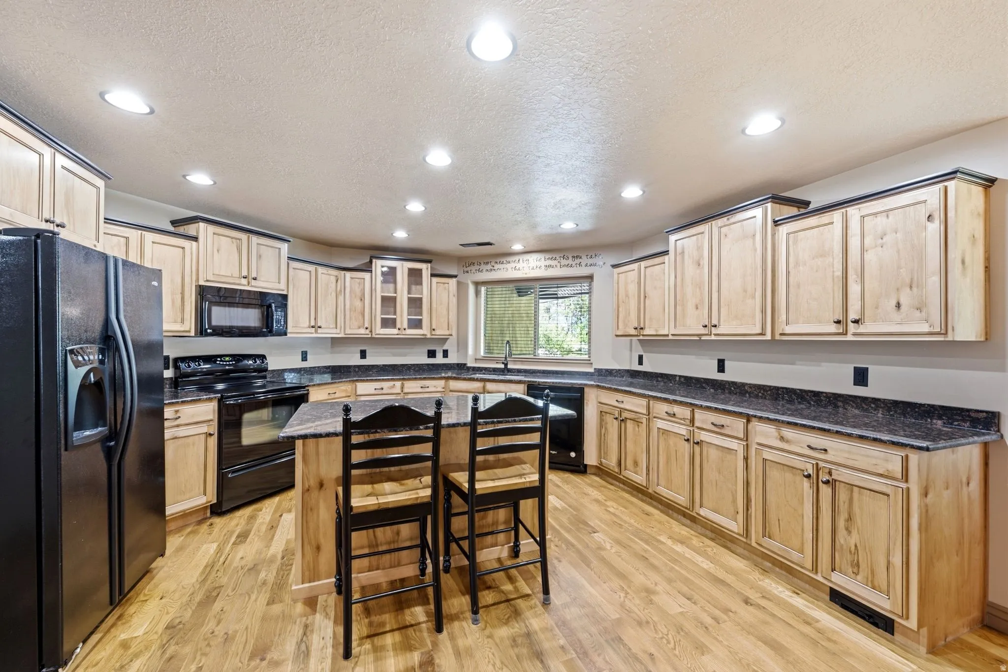 Expansive kitchen featuring light wood cabinetry with dark trim, black appliances, dark countertops, and a central island with bar seating