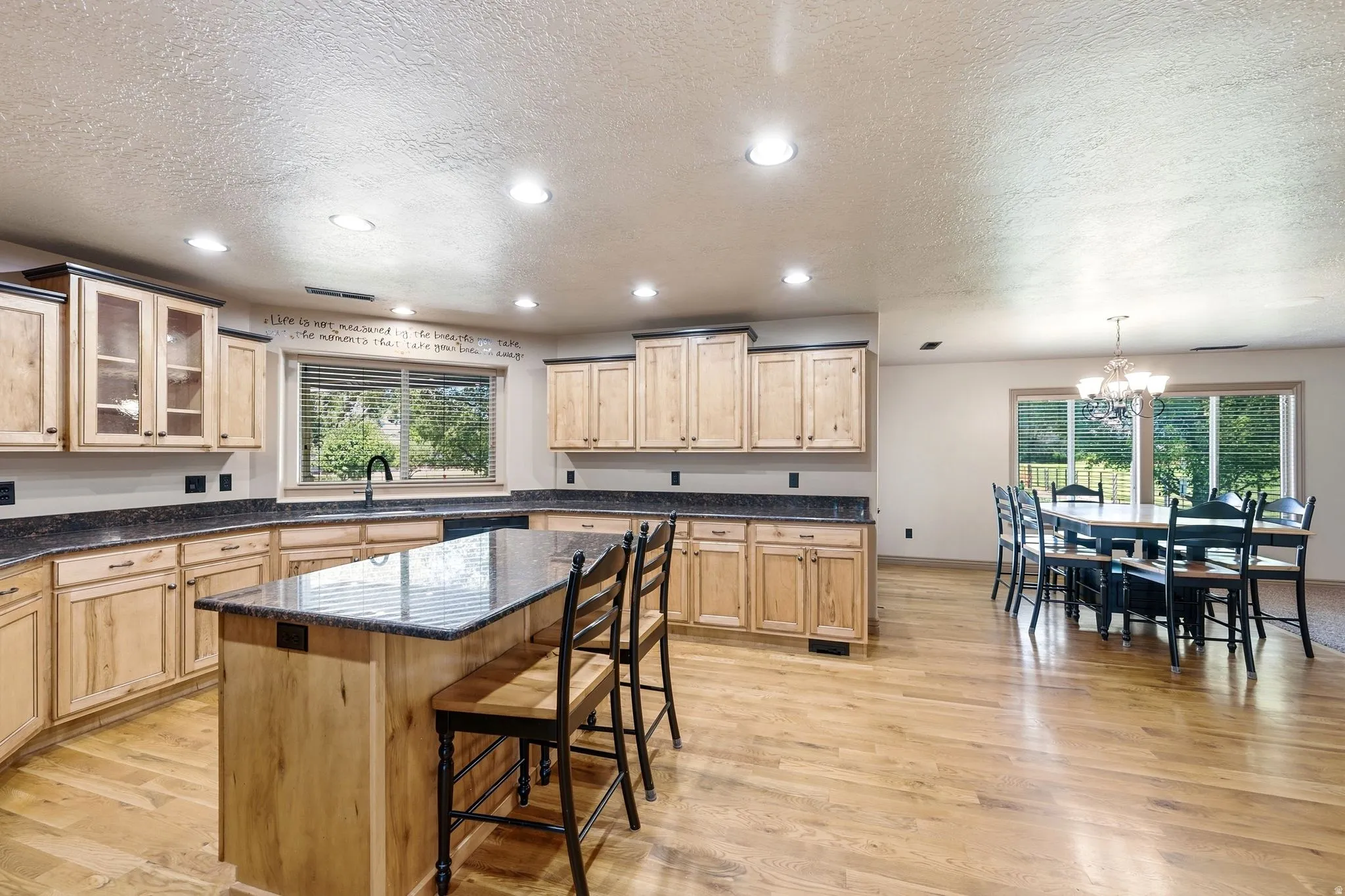 Open-concept kitchen featuring light wood cabinetry, dark countertops, a central island with bar seating, and wood-finish flooring