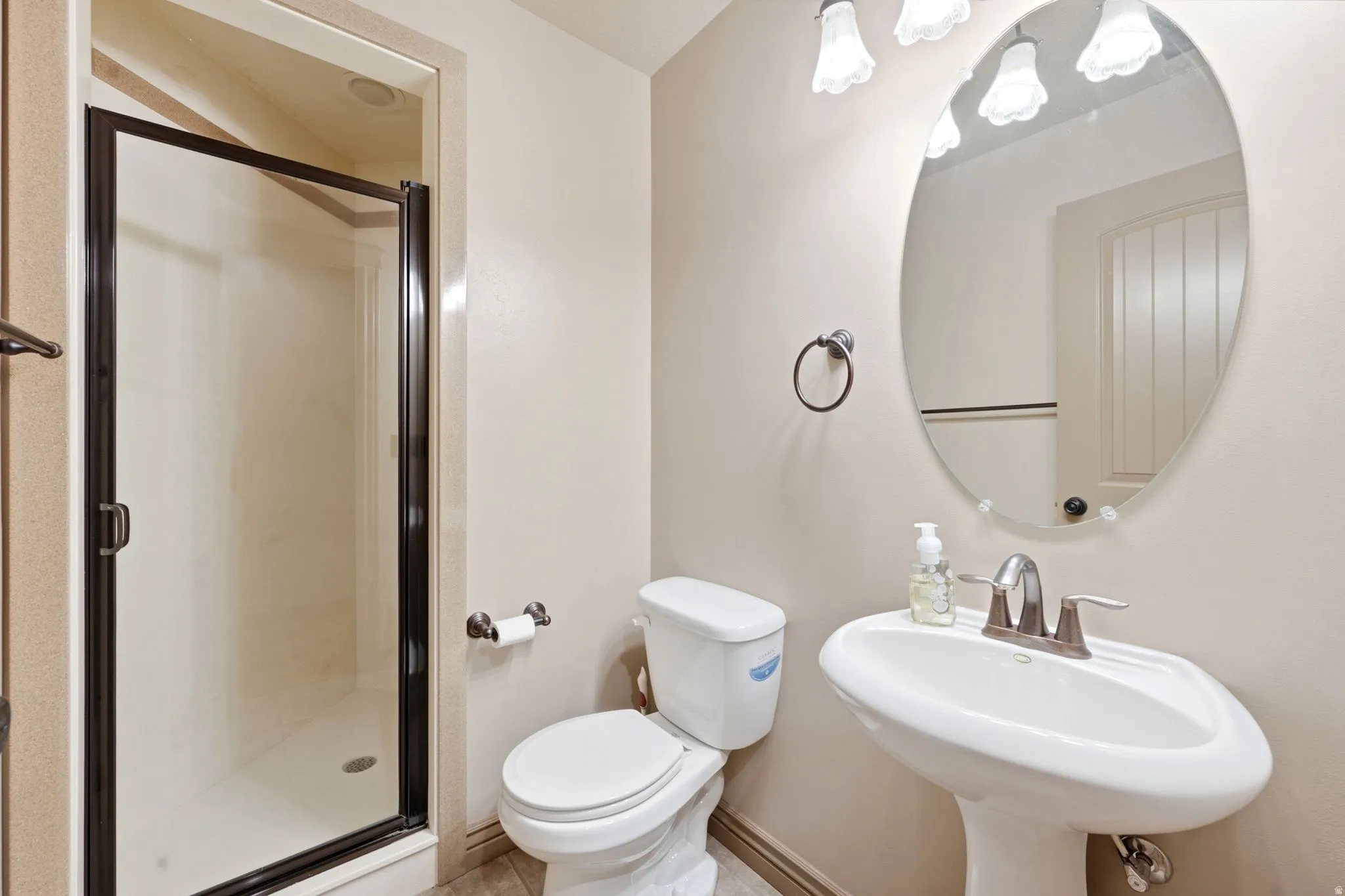 Bathroom featuring a pedestal sink, oval mirror, wall-mounted light fixture, toilet, and a stand-up shower with a dark-framed enclosure