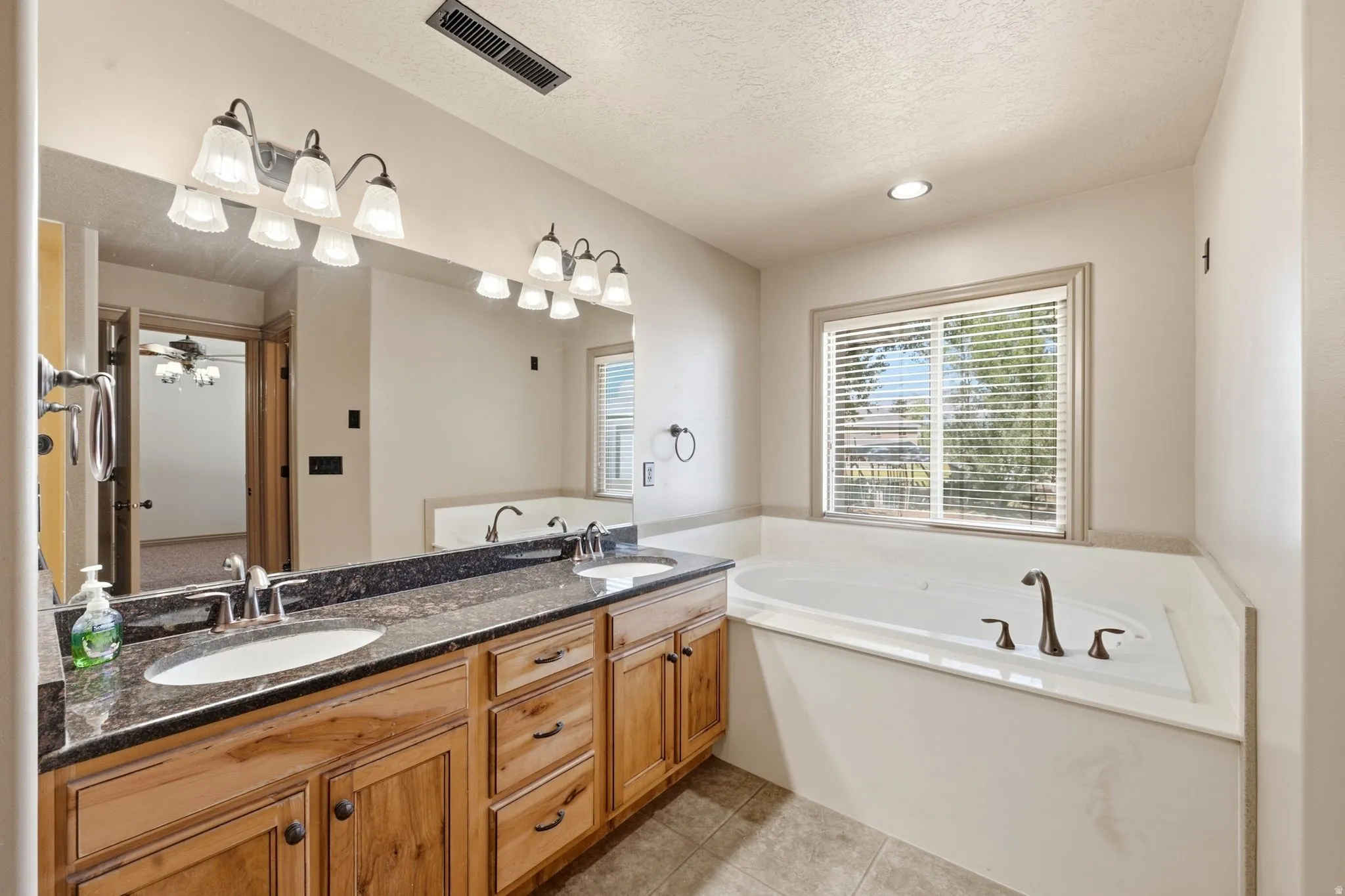 Bathroom featuring a spacious built-in tub with dark bronze fixtures, a dual sink vanity with a dark speckled countertop, light wood cabinetry, and overhead lighting