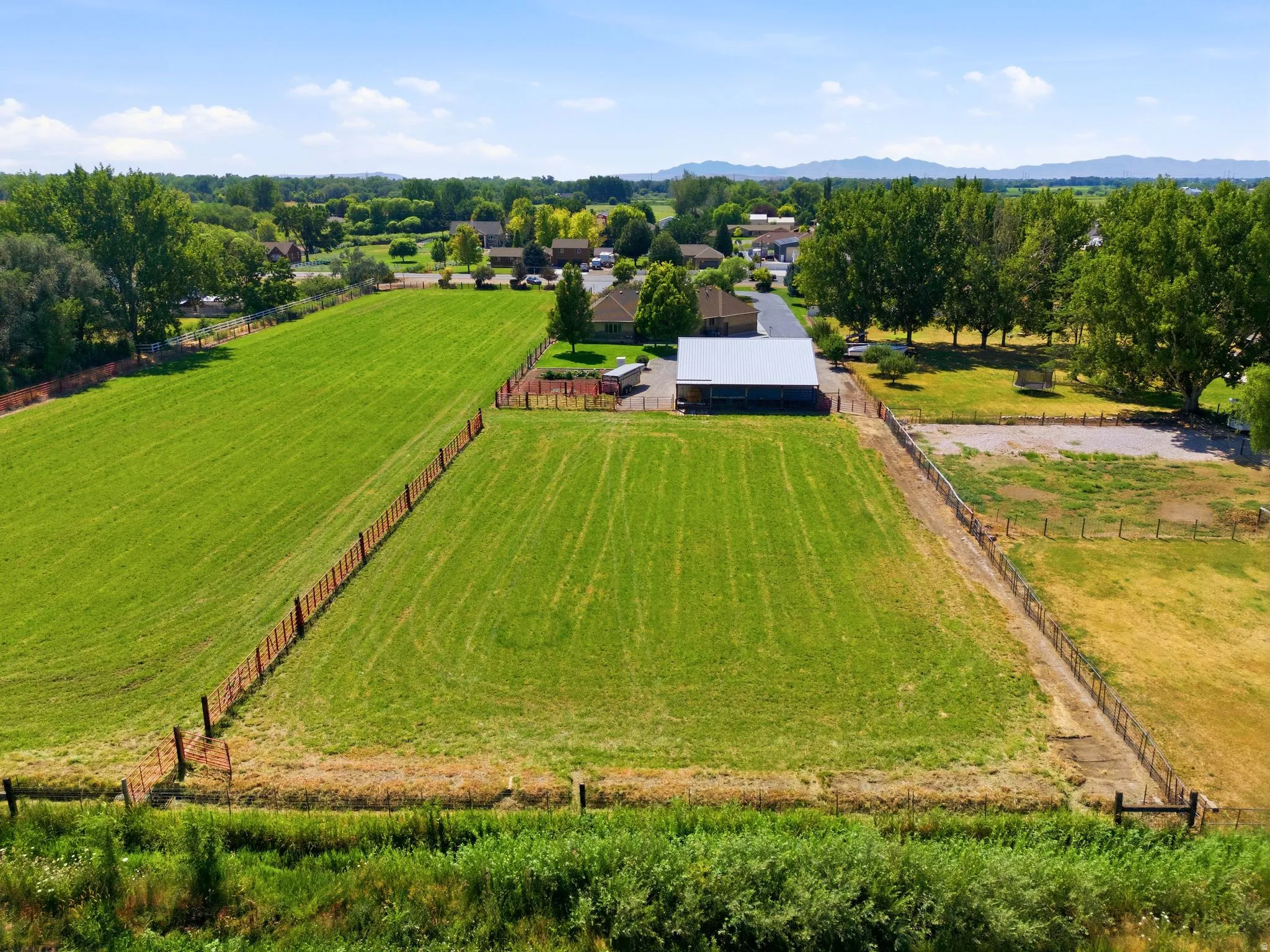 Expansive green fields featuring post and rail fencing, a large outbuilding, and mature trees