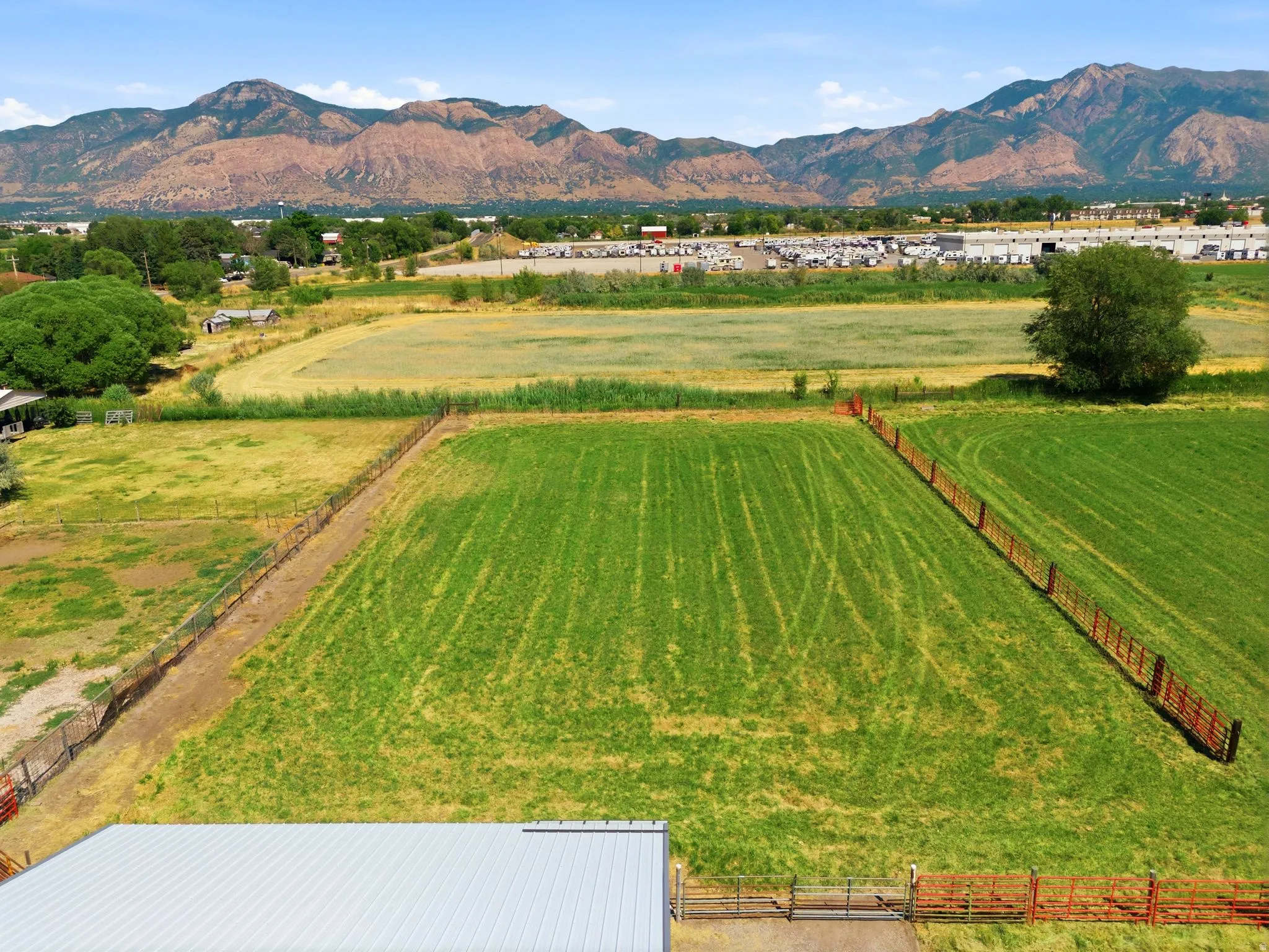 Expansive green field bordered by fencing and mature trees, featuring a distant mountainous backdrop and a visible metal-roof structure