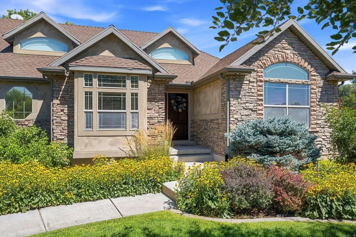 Stone facade home featuring a prominent bay window, arched window accents, dormer windows, a concrete pathway, and landscaped grounds with flowering plants