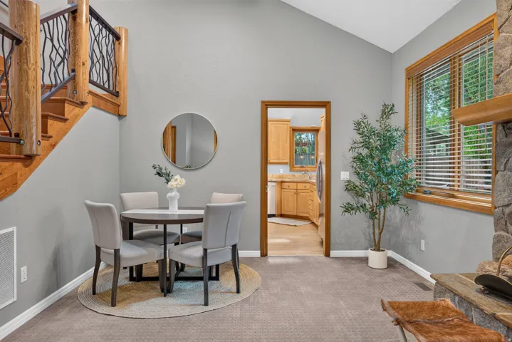 Vaulted ceiling with an open floor plan, a wood staircase featuring a decorative metal railing, a stone fireplace, wood-framed windows, and carpet flooring