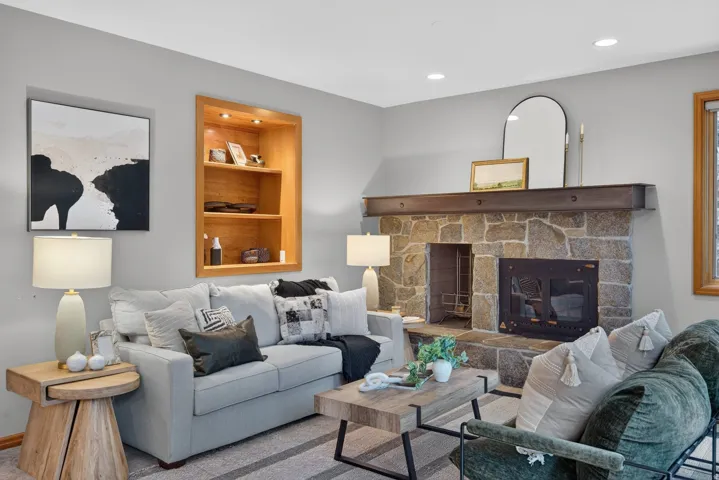 Living area featuring a stone-clad fireplace with a dark wood mantel, recessed lighting, a built-in illuminated wood shelving unit, light gray wall paint, and wood-trim windows