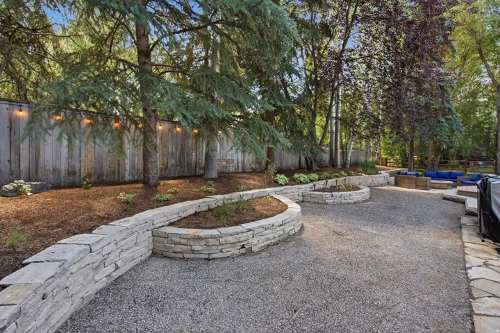 Landscaped backyard featuring layered stone retaining walls, a gravel patio area, mature trees, and a privacy fence with string lighting