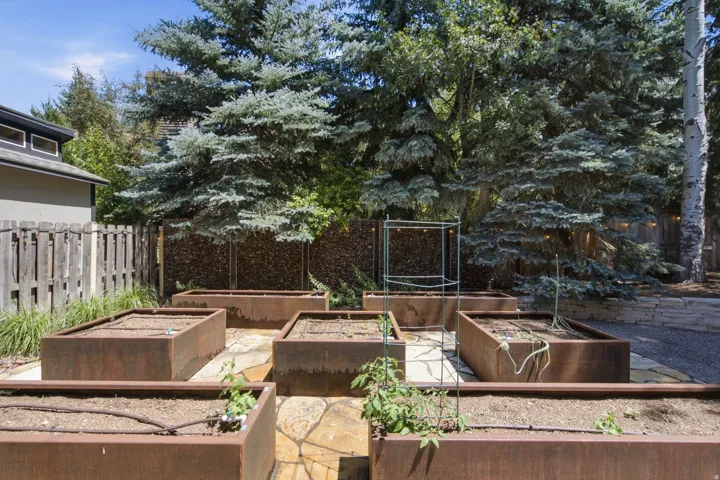 Garden area featuring multiple rectangular raised planters with drip irrigation, irregular flagstone paving, a gabion fence, and mature evergreen trees