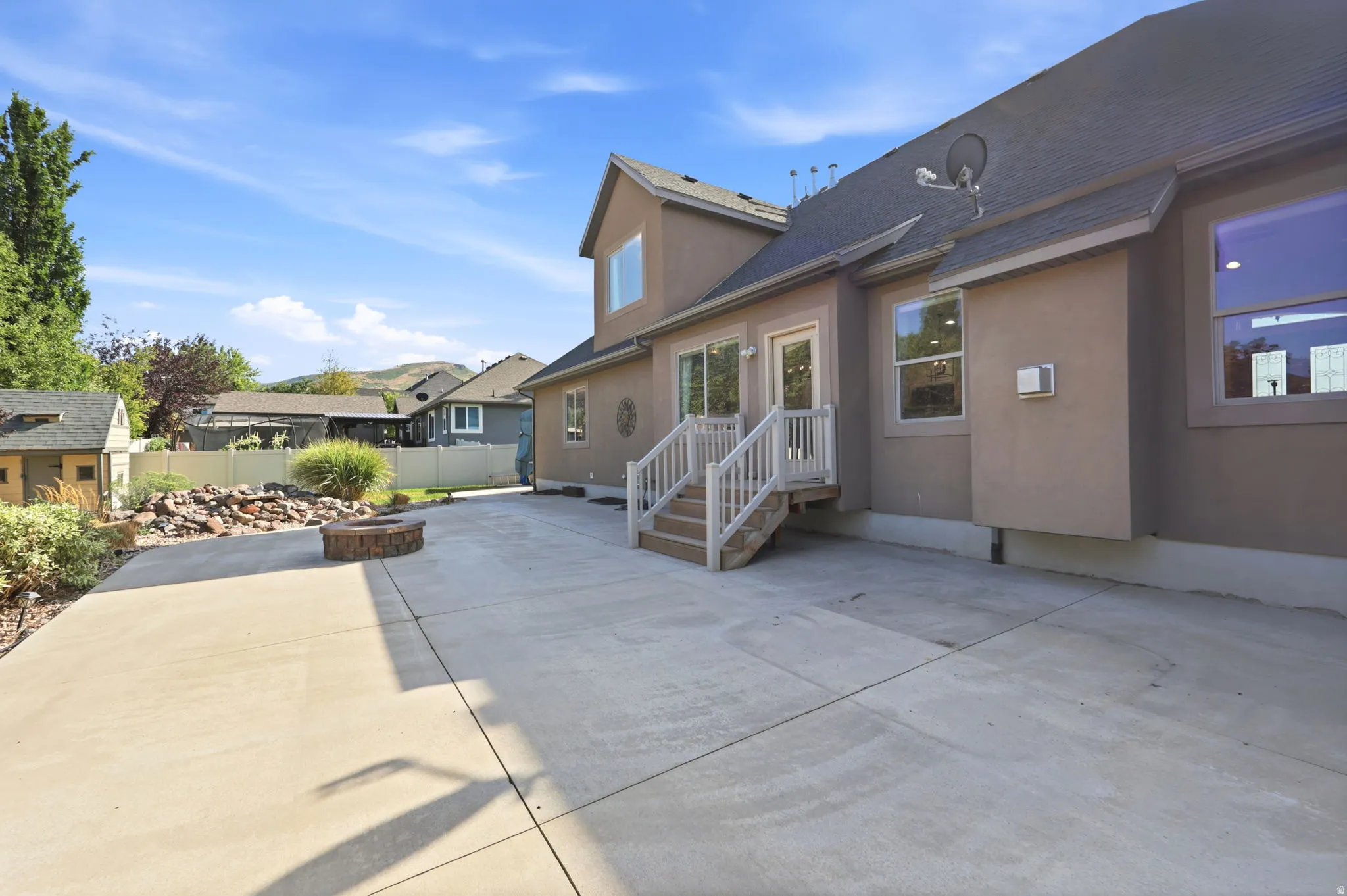 Expansive concrete patio featuring a built-in stone fire pit, stucco exterior, multiple window placements, and a rear entry with steps