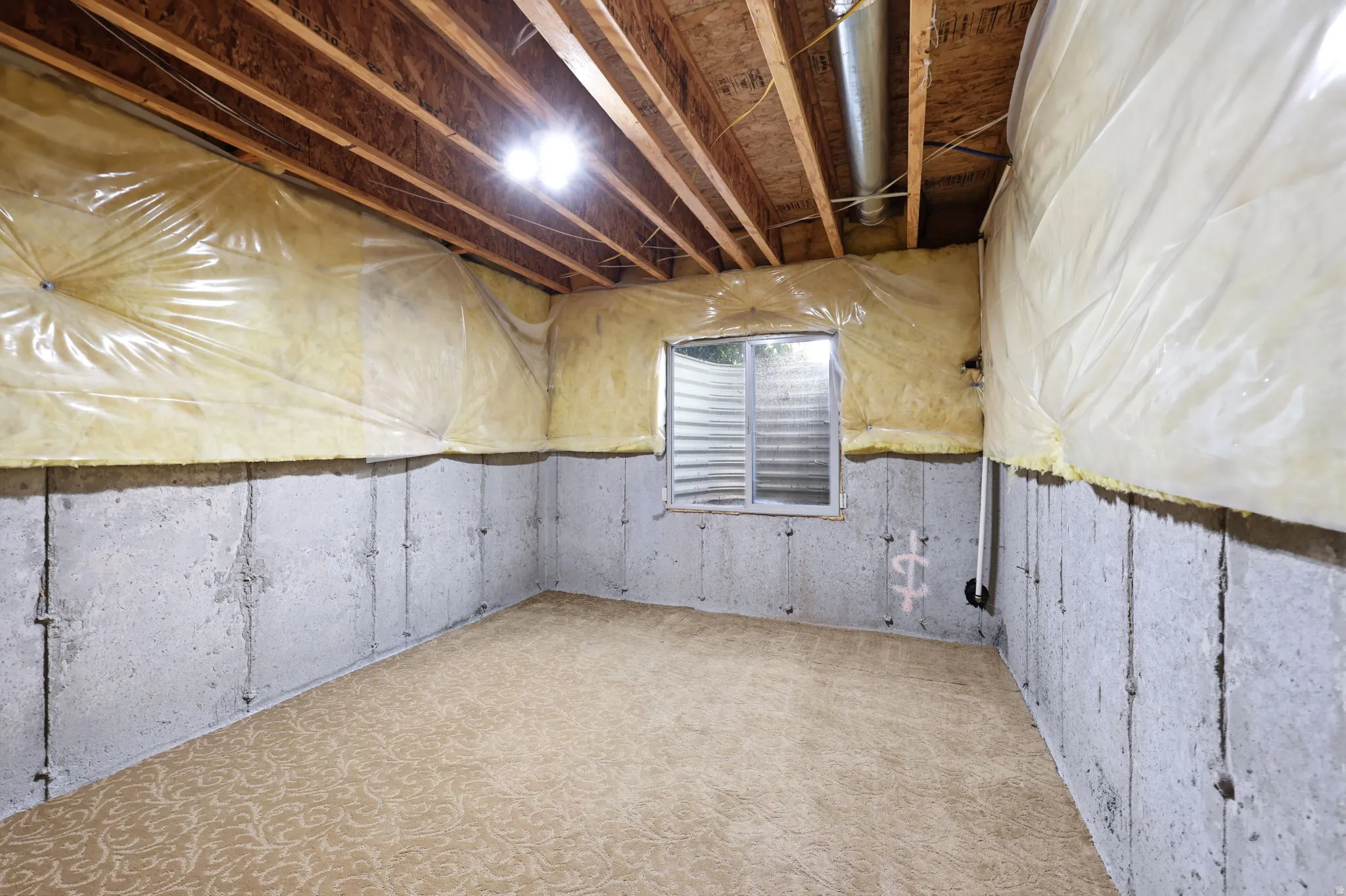 Unfinished basement featuring exposed wood joists, concrete foundation walls, a window well, and patterned carpeting
