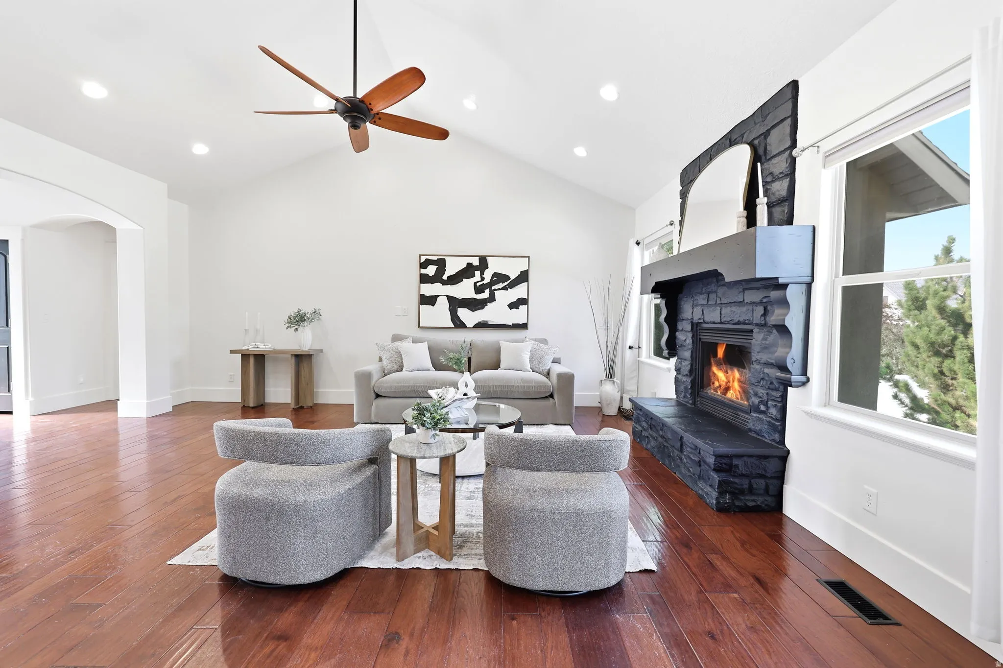 Spacious living area featuring high vaulted ceilings, rich wood-finish flooring, a large black stone fireplace with hearth, recessed lighting, and a ceiling fan