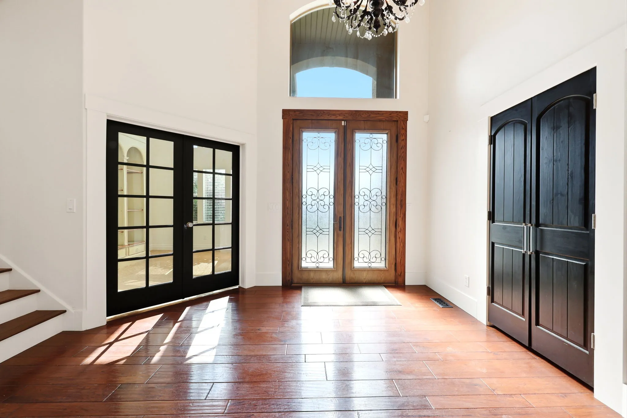 Grand entryway featuring wood-finish flooring, a double-door entry with decorative glass inserts and an arched transom window, a crystal chandelier, and black French doors
