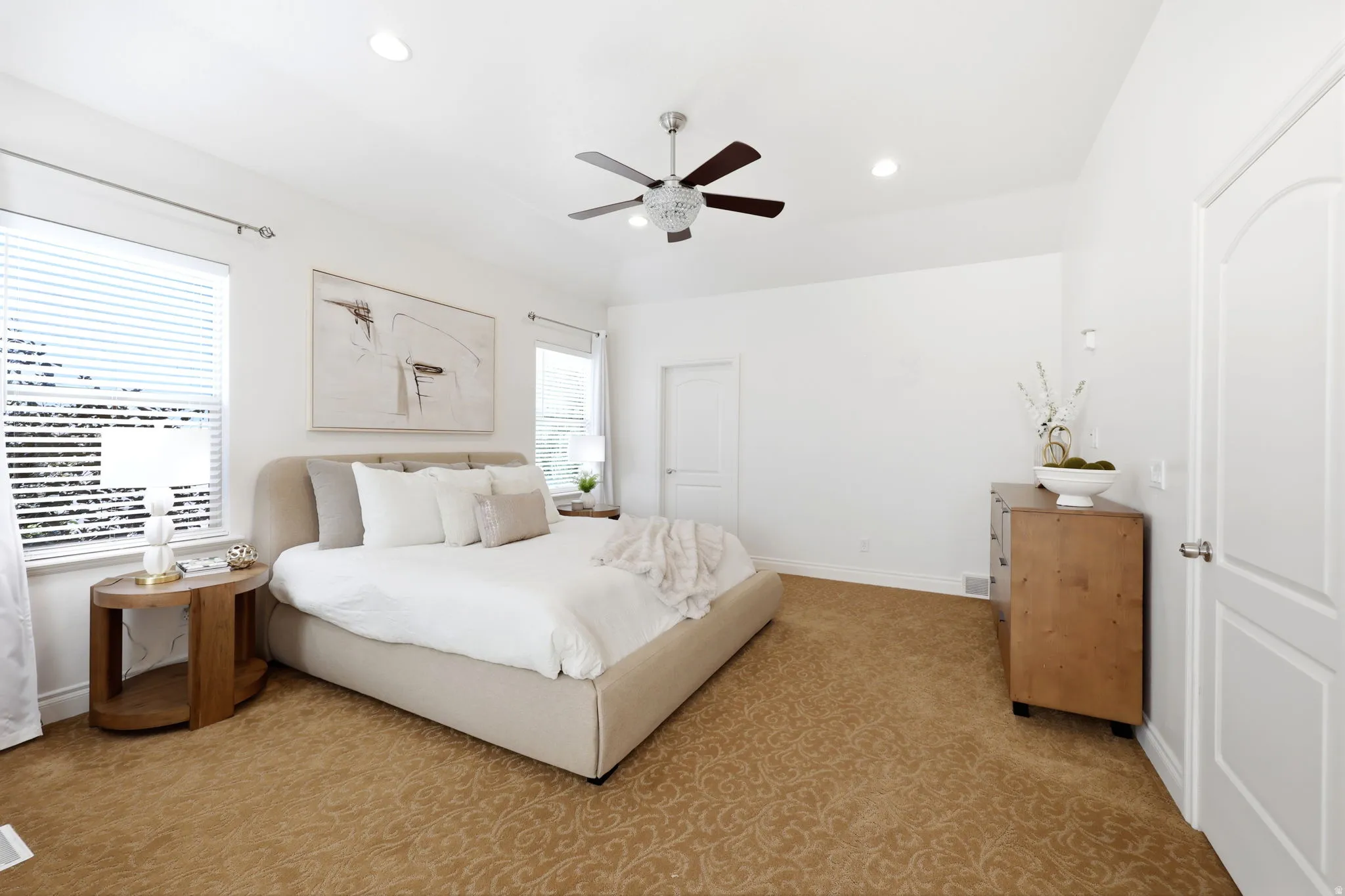 Spacious bedroom featuring light-colored walls, patterned carpeting, multiple windows with blinds, a ceiling fan with integrated lighting, and recessed lighting