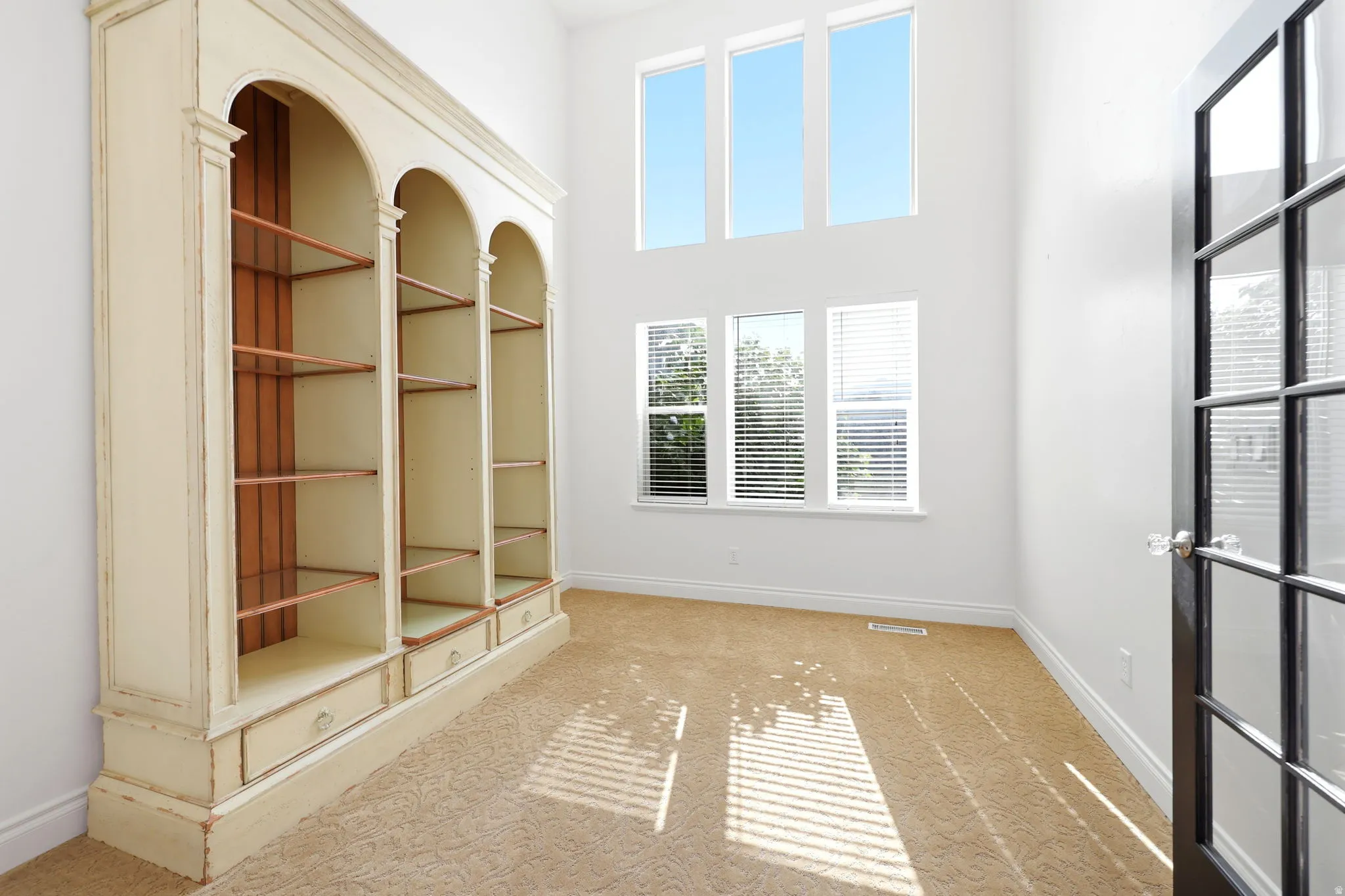 Room featuring a tall built-in bookshelf with arched details and integrated drawers, high ceilings, multiple windows with blinds, and a glass-paned door