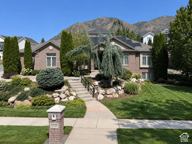 Brick-clad exterior with a prominent entry staircase, mature landscaping, integrated solar panels, and a manicured lawn