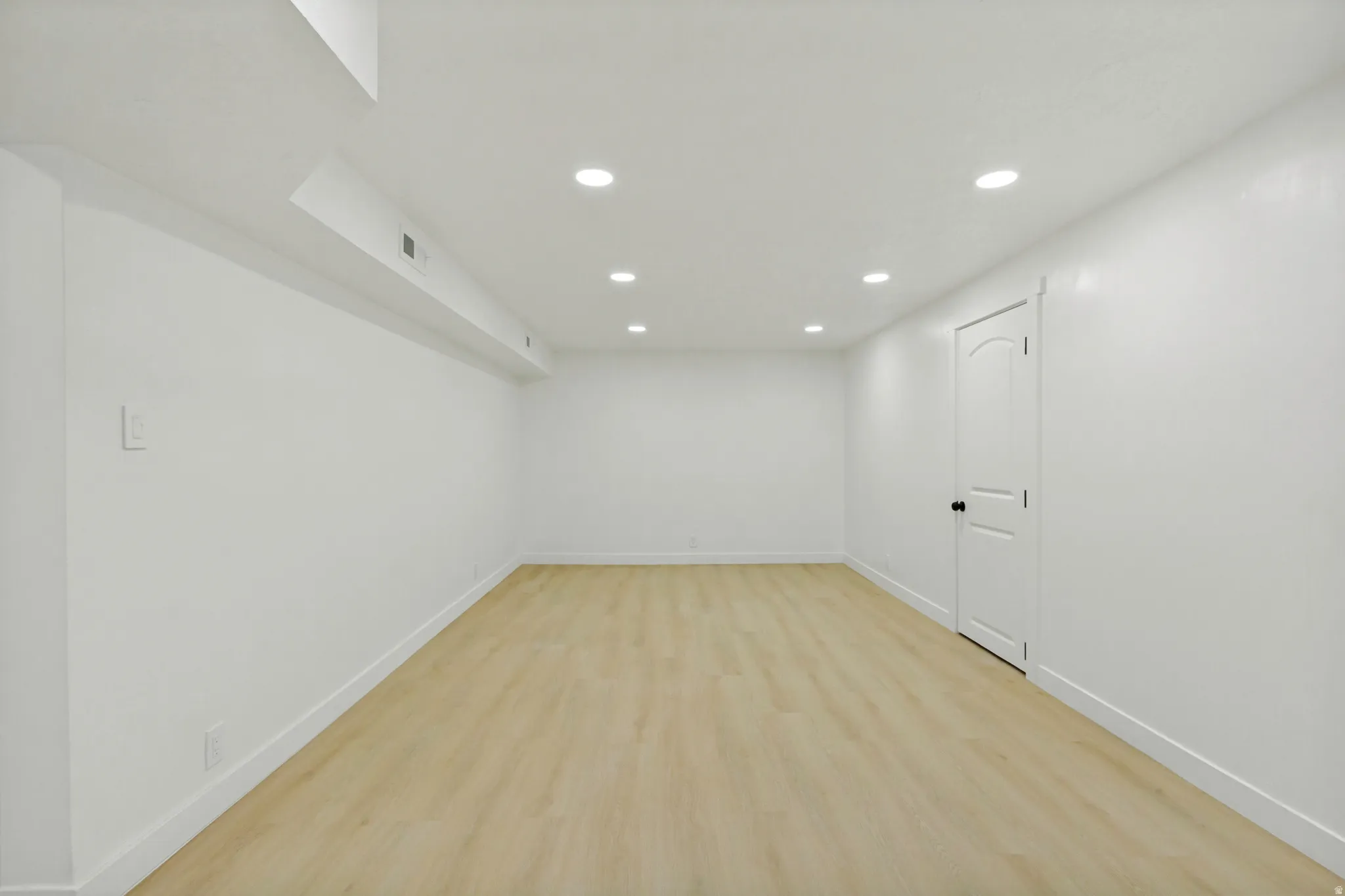 Spacious room featuring light wood-finish flooring, white walls, recessed ceiling lighting, and a solid white door with black hardware
