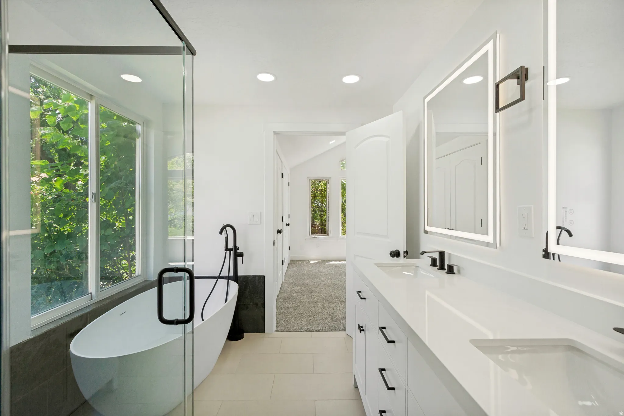 Bathroom featuring a freestanding soaking tub, a dual vanity with integrated sinks, back-lit mirrors, matte black fixtures, and a glass-enclosed shower