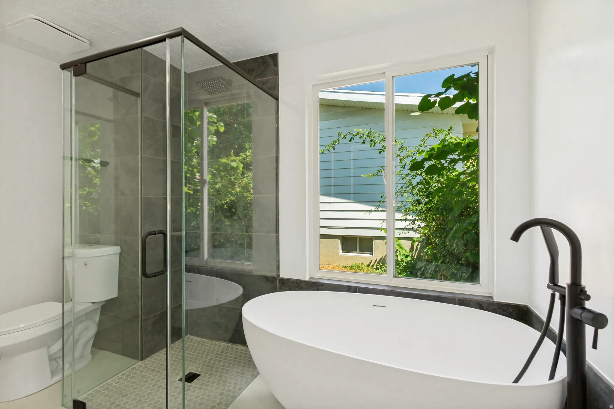 Freestanding oval soaking tub with a matte black floor-mounted faucet, complemented by a glass-enclosed shower with gray tiled walls and hexagonal mosaic flooring