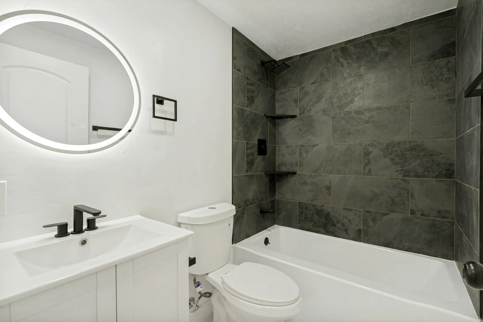 Bathroom featuring a white vanity with integrated sink, matte black fixtures, an illuminated circular mirror, and a shower-over-tub with dark large-format tile surround and built-in shelving