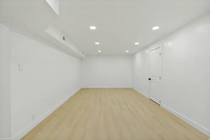 Spacious room featuring light wood-finish flooring, white walls, recessed ceiling lighting, and a solid white door with black hardware