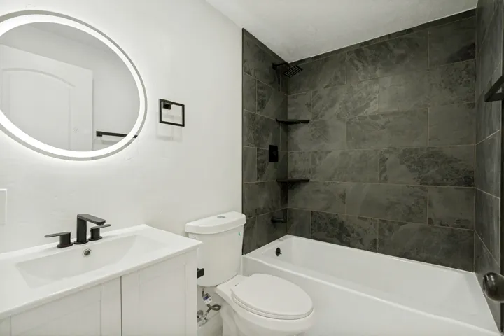 Bathroom featuring a white vanity with integrated sink, matte black fixtures, an illuminated circular mirror, and a shower-over-tub with dark large-format tile surround and built-in shelving