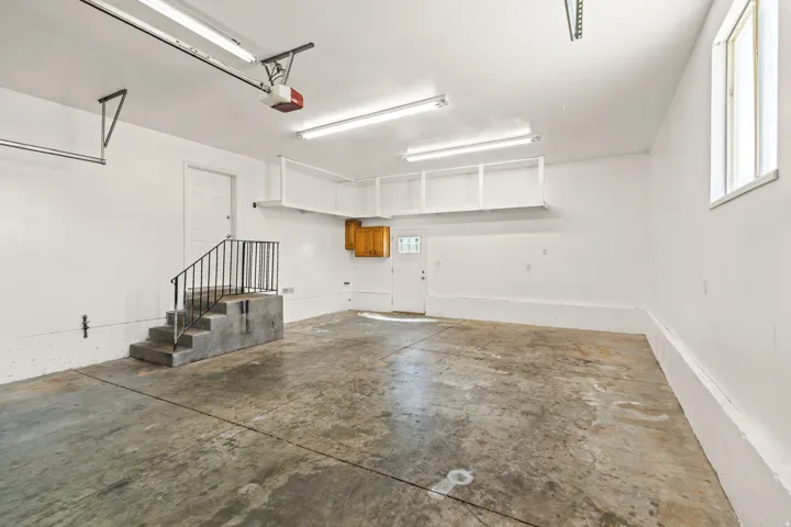 Spacious garage featuring a concrete floor, bright overhead lighting, a single window, built-in overhead shelving, and an interior door with steps and a metal handrail