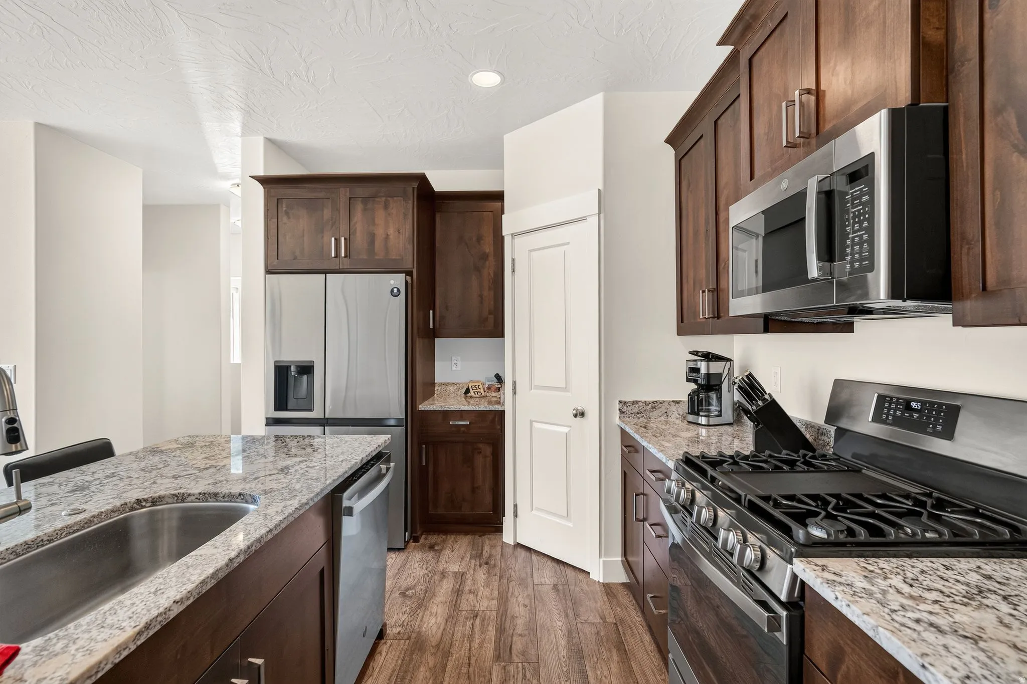Modern kitchen featuring dark wood cabinetry, light granite countertops, stainless steel appliances, and wood-finish flooring
