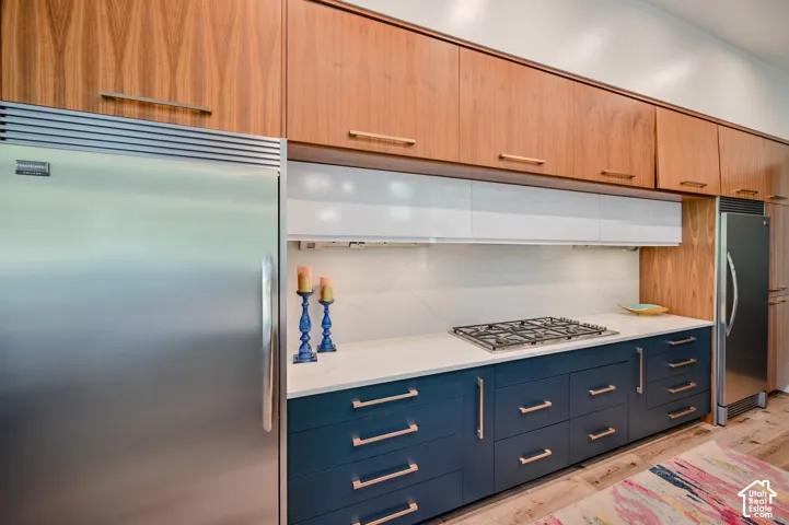 Contemporary kitchen featuring two-tone cabinetry with wood-finish upper cabinets and deep blue lower cabinets, stainless steel appliances, a built-in gas cooktop, and a light-toned countertop with a matching backsplash