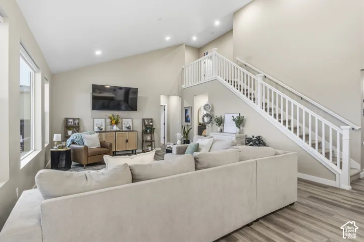 Spacious living area featuring a vaulted ceiling with recessed lighting, wood-finish flooring, multiple large windows, and a prominent staircase with white railings