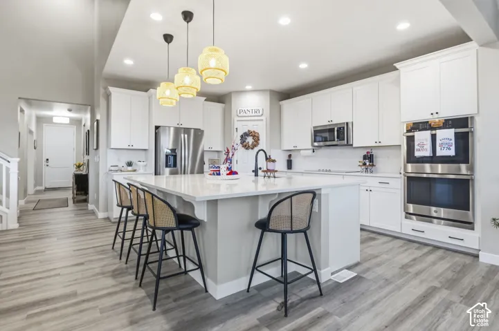 Open-concept kitchen featuring an oversized island with seating, pendant lighting, white shaker cabinetry, stainless steel appliances, and wood-finish flooring