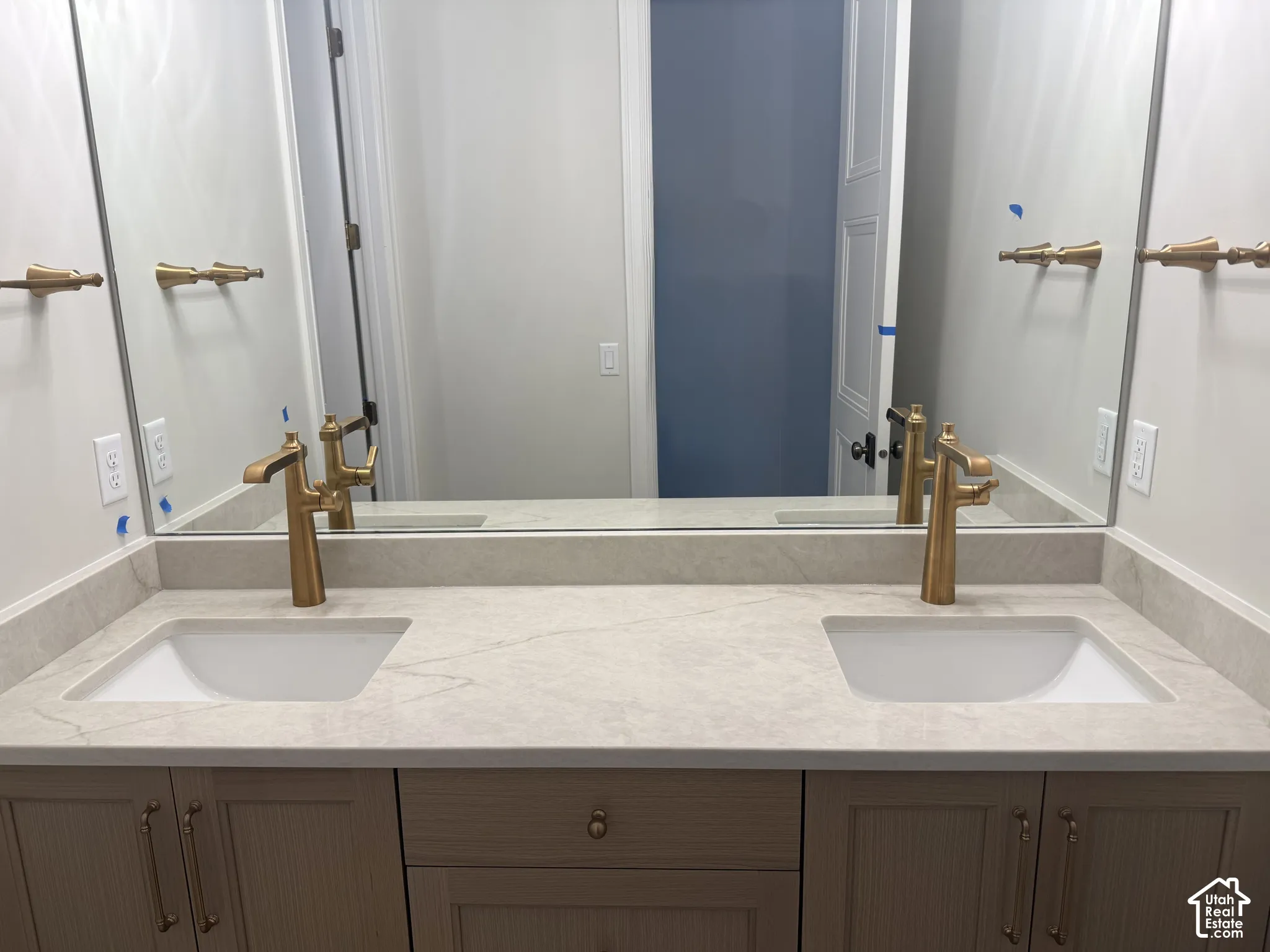 Double vanity with a stone countertop, under-mount sinks, brass-toned fixtures, and light-toned shaker-style cabinetry topped with a full-width mirror