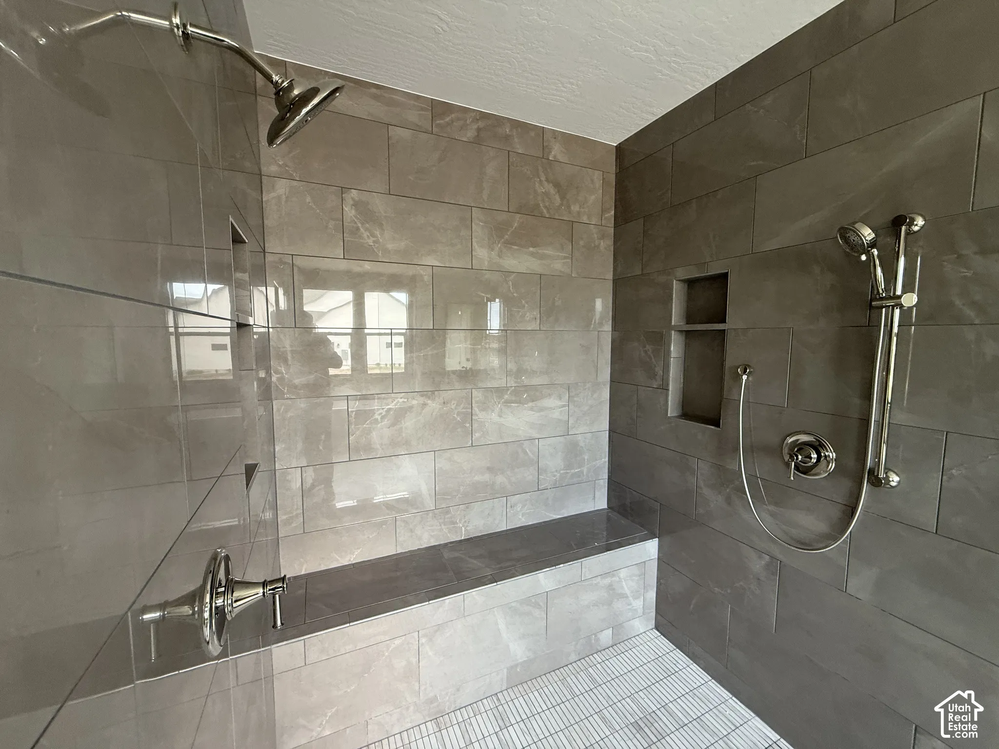 Walk-in shower featuring large-format ceramic wall tile, built-in tiled bench, dual showerheads with a handheld attachment, and recessed wall shelving