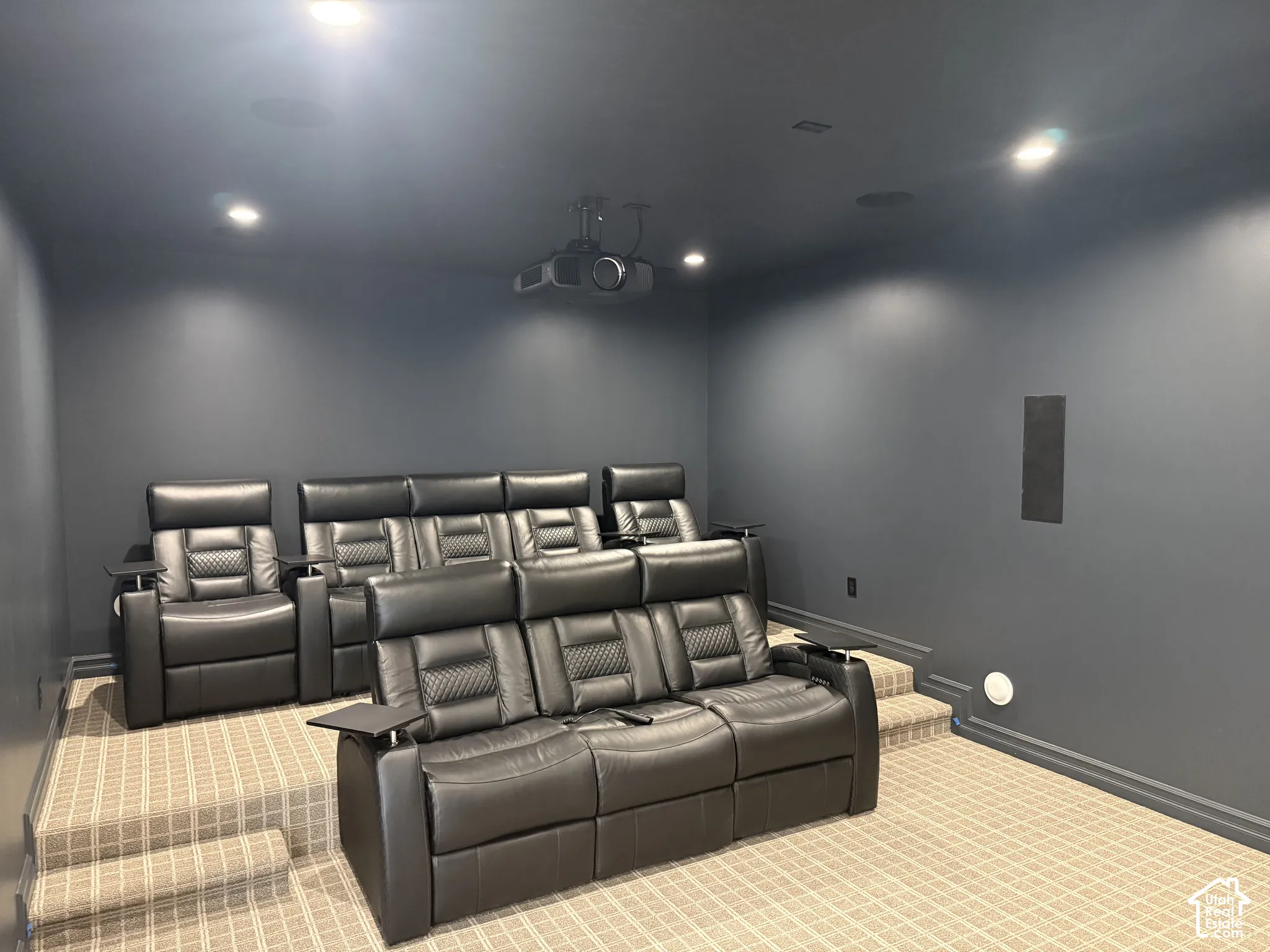 Dedicated media room featuring tiered carpeted flooring, built-in ceiling projector, recessed lighting, and integrated wall-mounted speakers