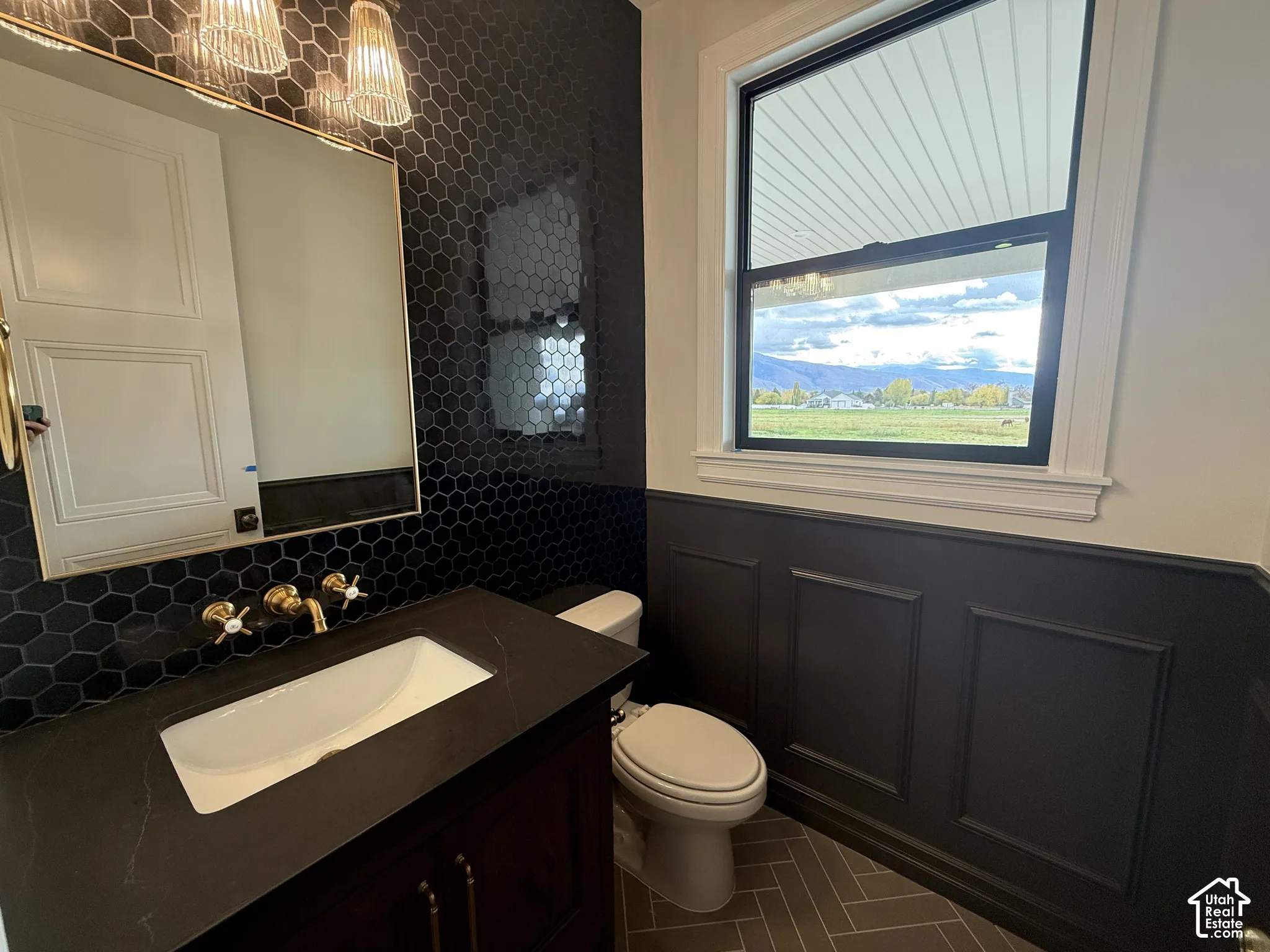Black hexagon tile backsplash, dark-toned vanity with a white undermount sink, brass wall-mounted faucet, wainscoting detail, and herringbone-patterned floor tiling