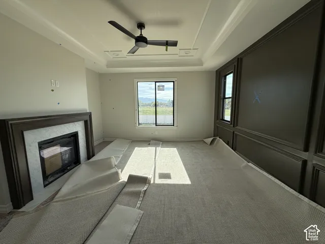 Tray ceiling with crown molding, ceiling fan, fireplace with a wood-finish mantel and stone surround, two windows, and a paneled accent wall