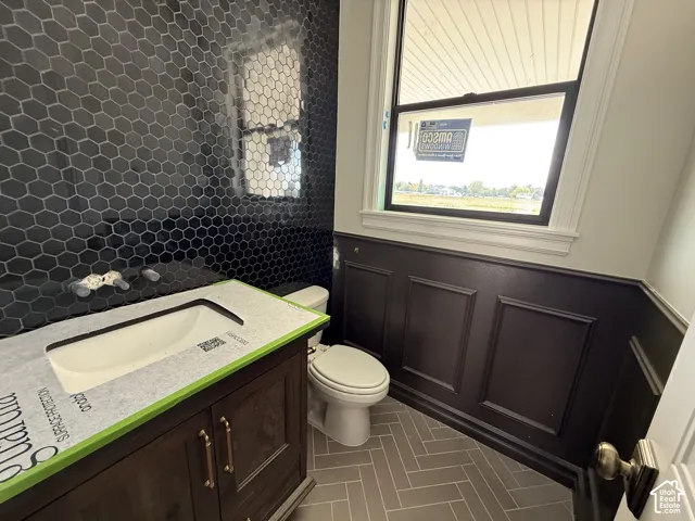 Dark hexagon tile accent wall, wood-finish vanity with integrated sink, panel wainscoting, and herringbone pattern floor tile
