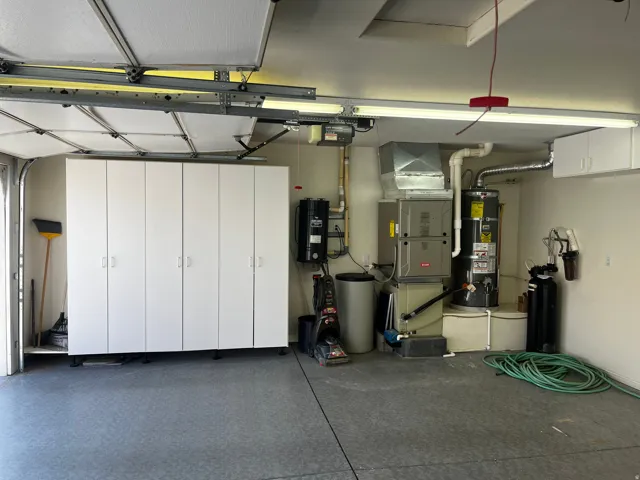 Garage featuring extensive built-in white cabinetry, textured flooring, overhead fluorescent lighting, and accessible utility systems