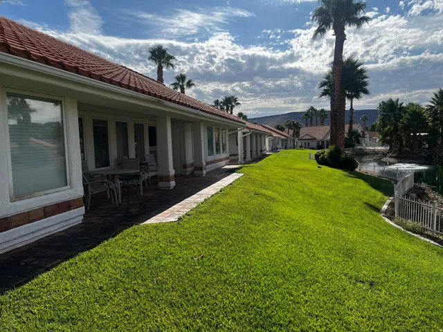 Expansive green lawn bordering a private patio with a tile roof, mature palm trees, and a decorative pond with a white fence