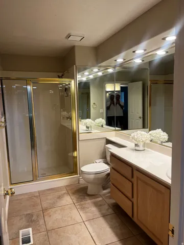 Full bathroom featuring a glass-enclosed shower with gold-tone framing, a double vanity with light wood-finish cabinetry and a white countertop, and large mirrored walls with integrated lighting