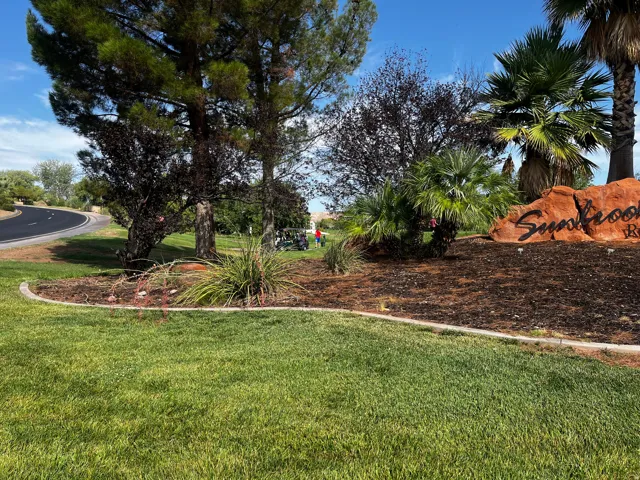 Landscaped entrance featuring a large stone sign, mature trees, palm trees, and lush green grass
