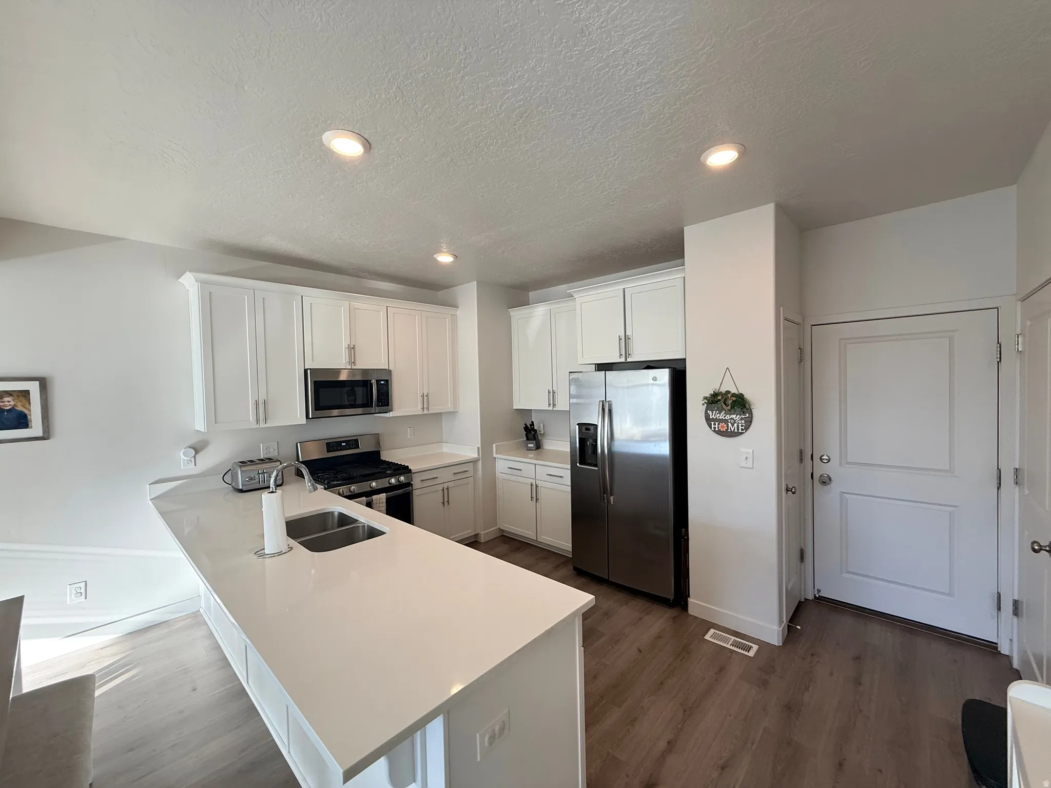 Bright kitchen featuring white shaker cabinetry, stainless steel appliances, a center island with an undermount dual-basin sink, light-toned countertops, and wood-finish flooring